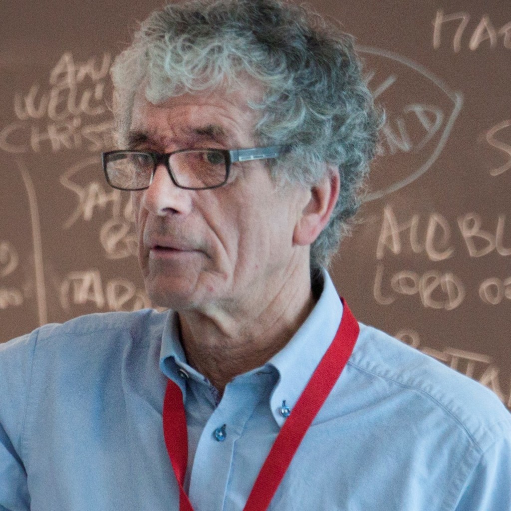 Brian Trench wearing a blue shirt and red lanyard standing in front of a blackboard with writing on it.