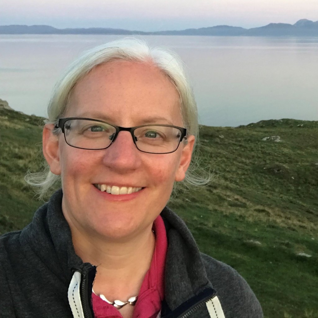 Jenn Dunbar with red top and black jacket standing outside with the sea in the background