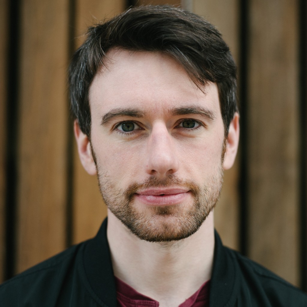 Alan Bennett wearing a black jacket standing in front of a wooden wall